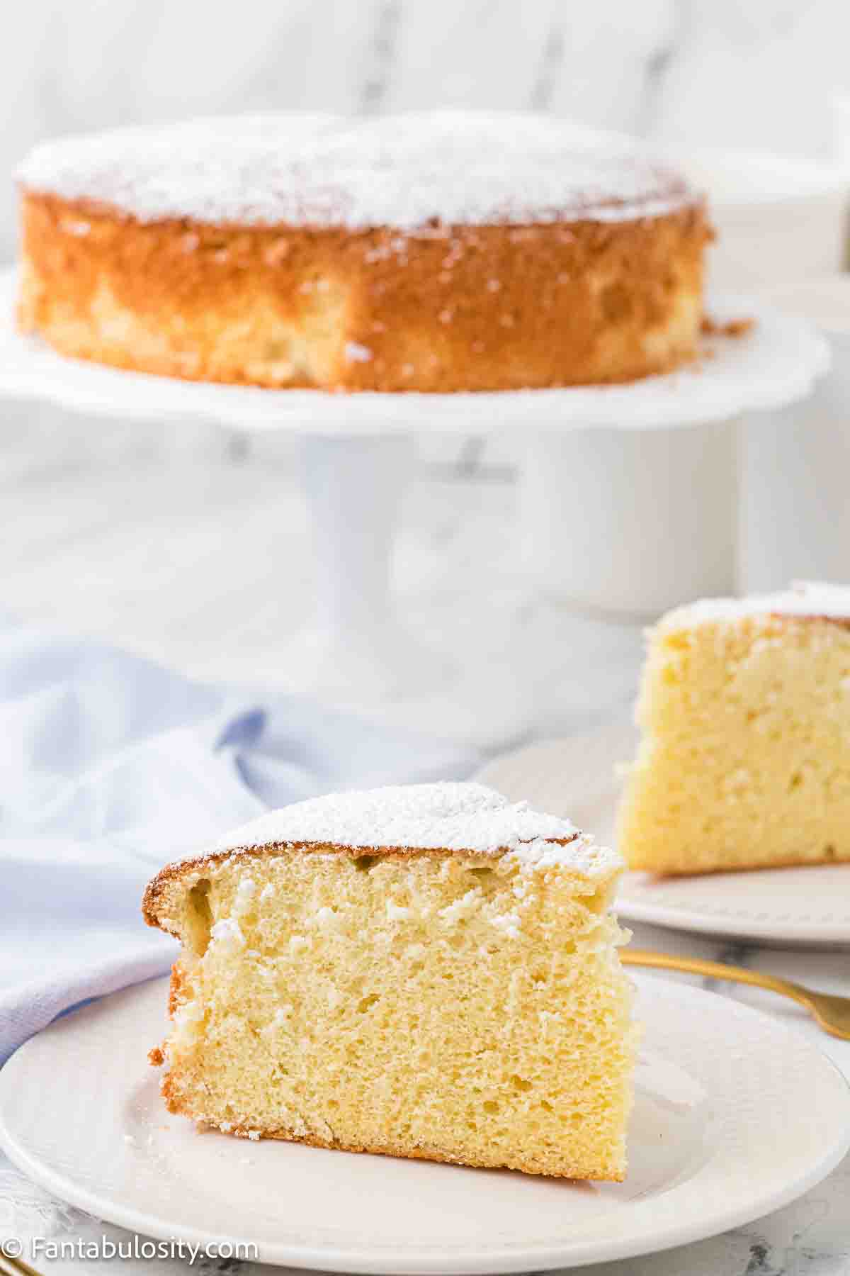 A slice of cloud cake on a round plate, sitting next to cake stand of rest of cake.