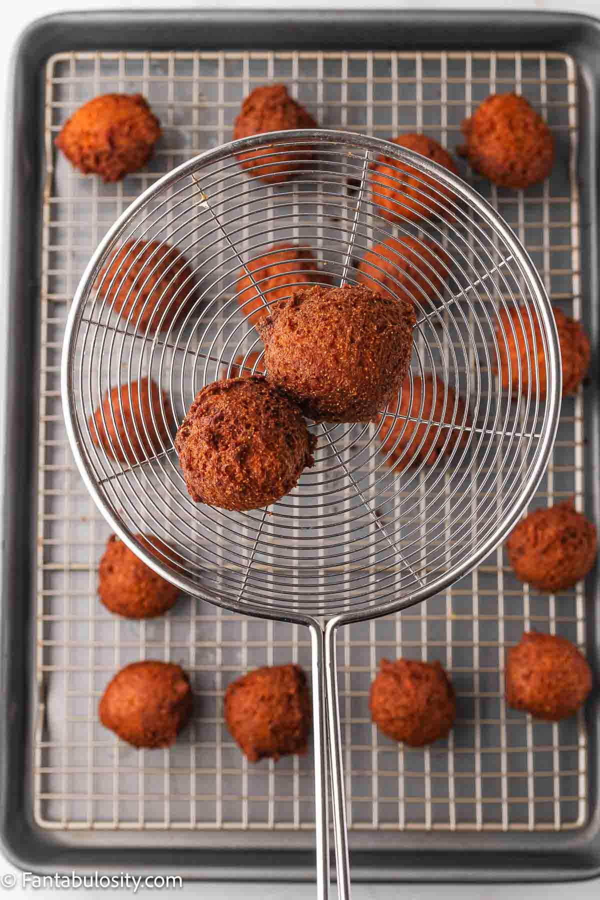 Cooked hush puppies in strainer spoon, resting above a wire rack where more hush puppies are sitting.