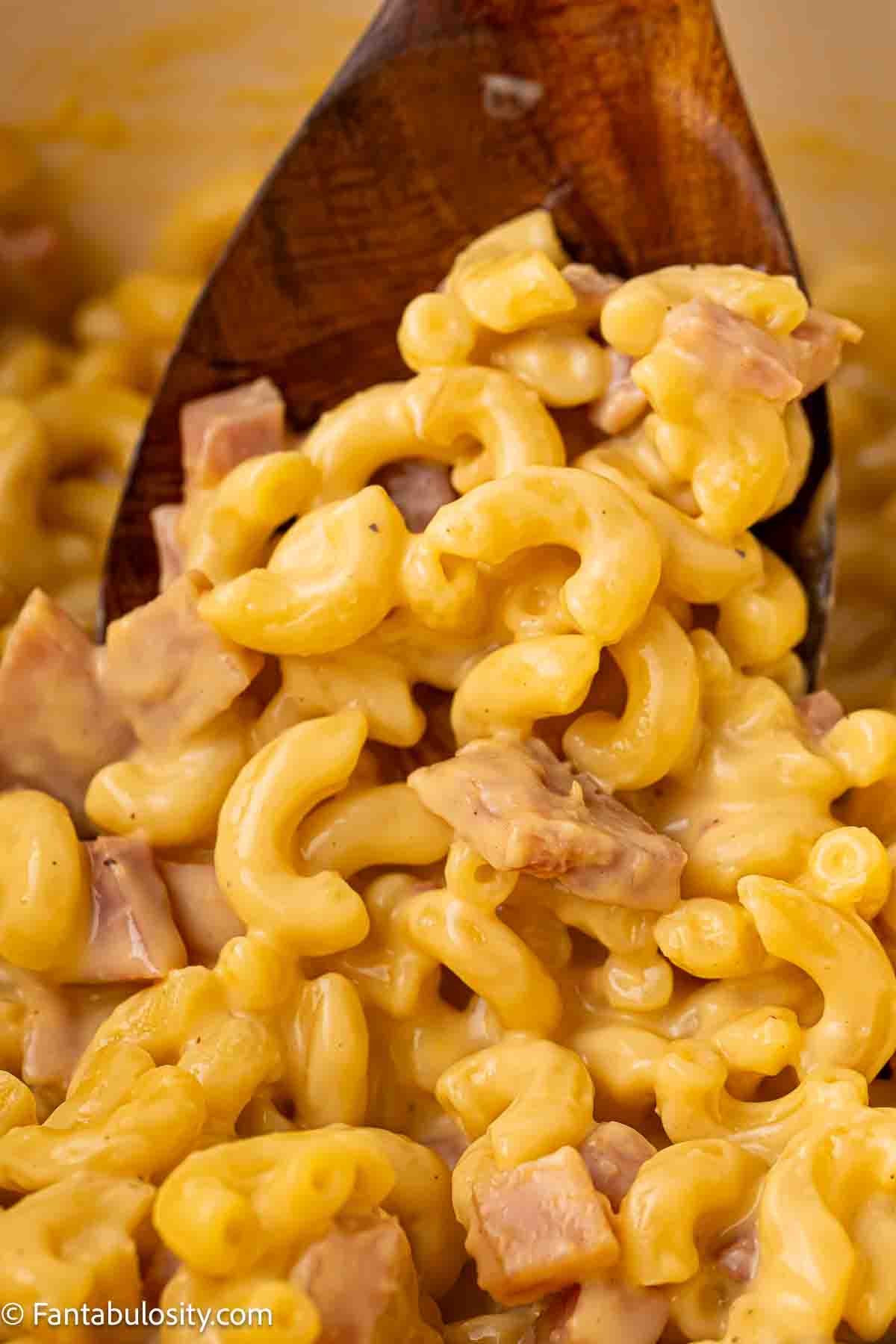 A wooden spoon sticking in Mac and Cheese with Ham.