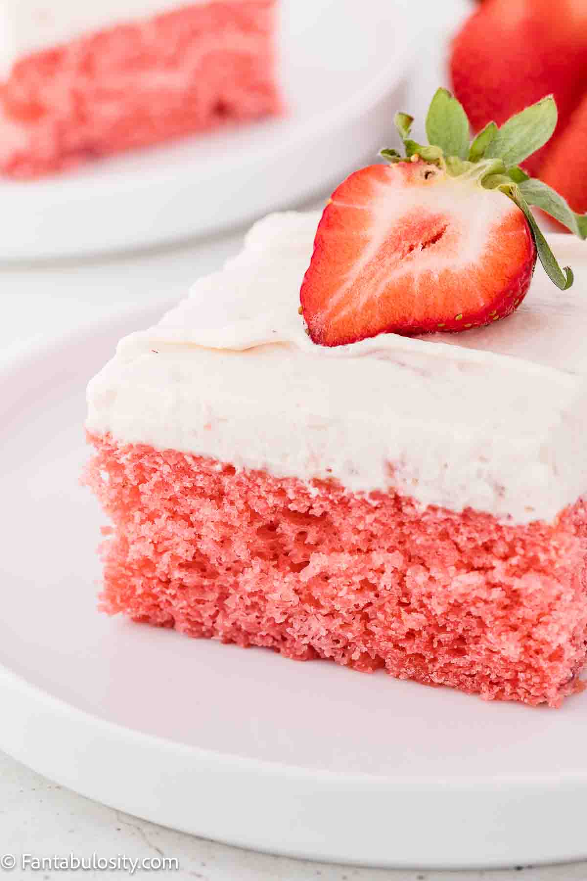 A slice of strawberry jello cake, topped with a strawberry, sitting on a round plate.