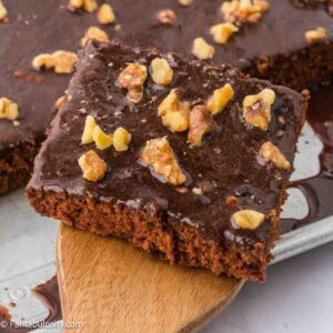 A slice of Texas sheet cake topped with walnuts, sitting on a wooden spatula, next to the rest of the pan.