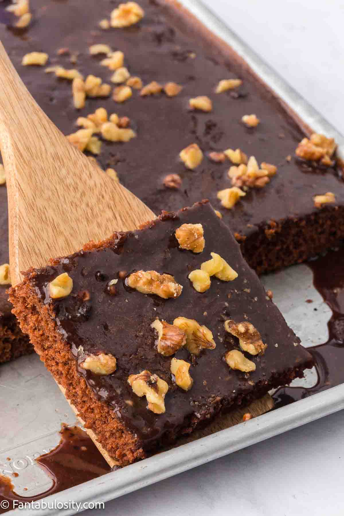 A slice of Texas Sheet Cake in baking pan, with a piece lifted out with a wooden spoon.