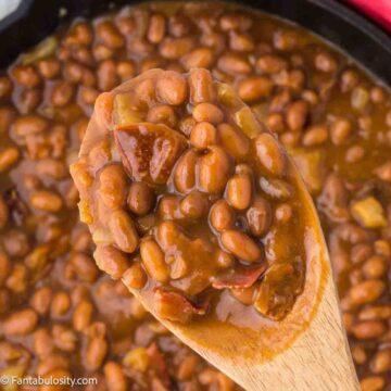 Dr. Pepper baked beans in a cast iron and wooden spoon spooning out a serving.