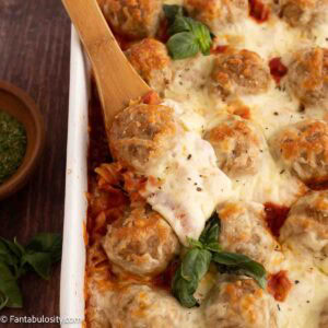 A wooden spoon dishing out a serving of meatball casserole in a 9x13 baking dish.