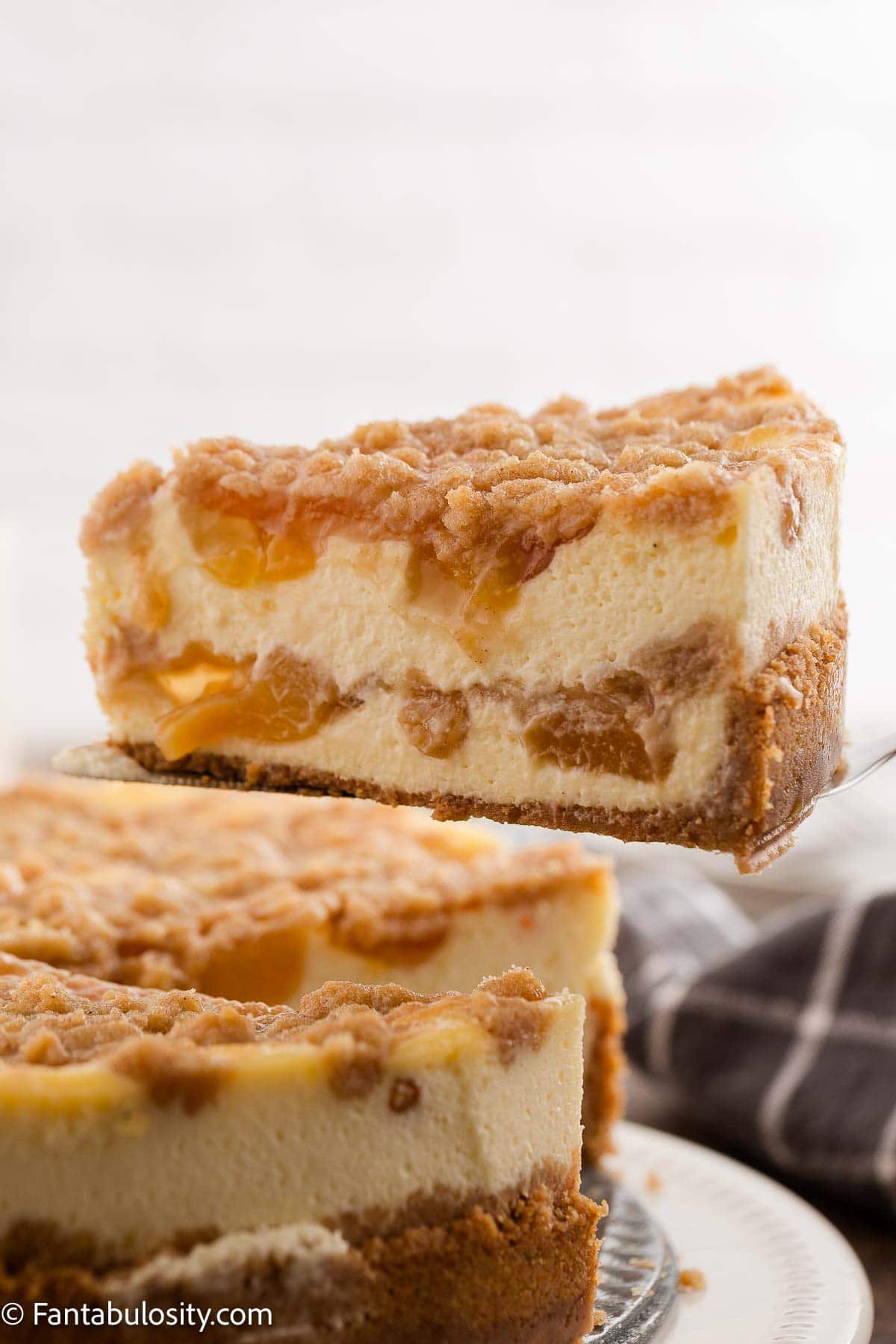 A slice of peach cobbler cheesecake on a spatula, next to rest of cheesecake.