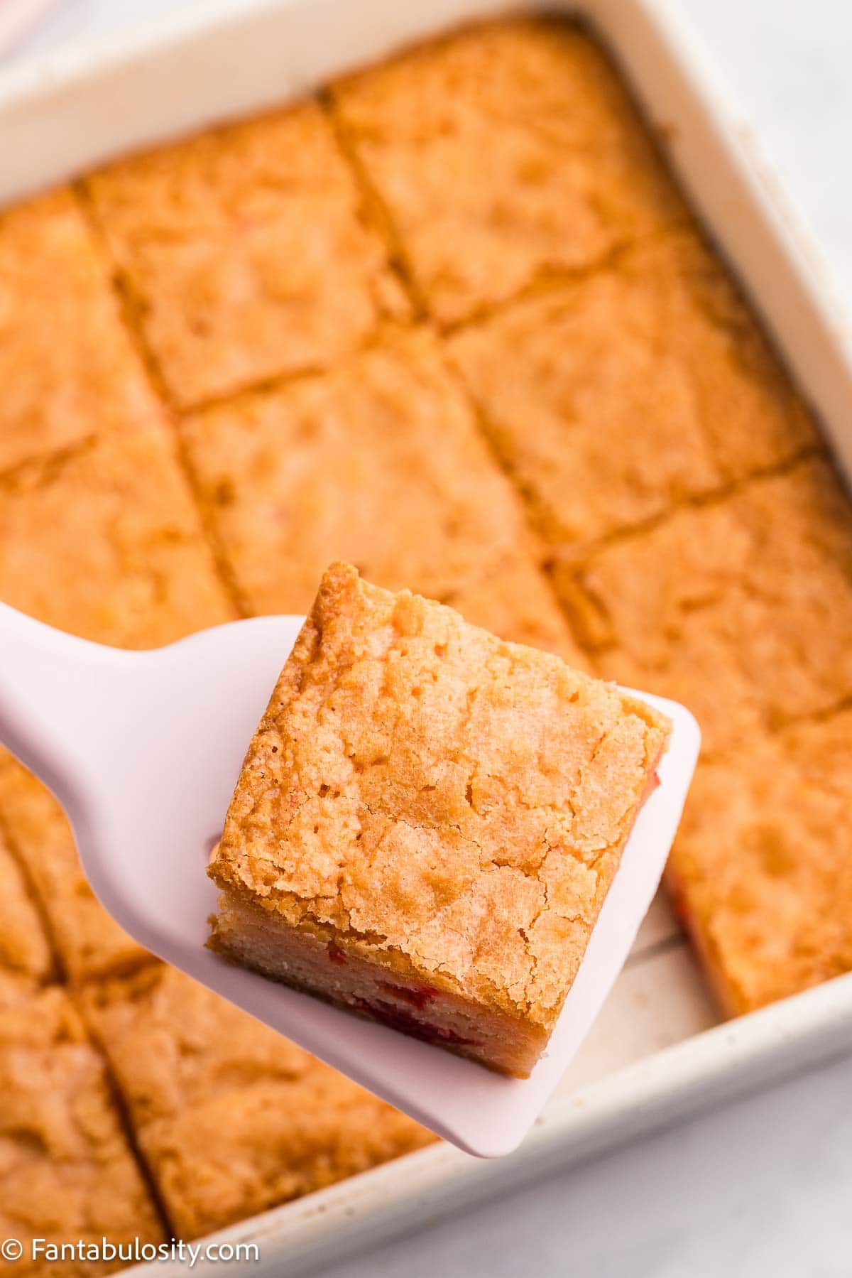 Baked cherry blondies in 8x8 baking pan with spatula holding one blondie.