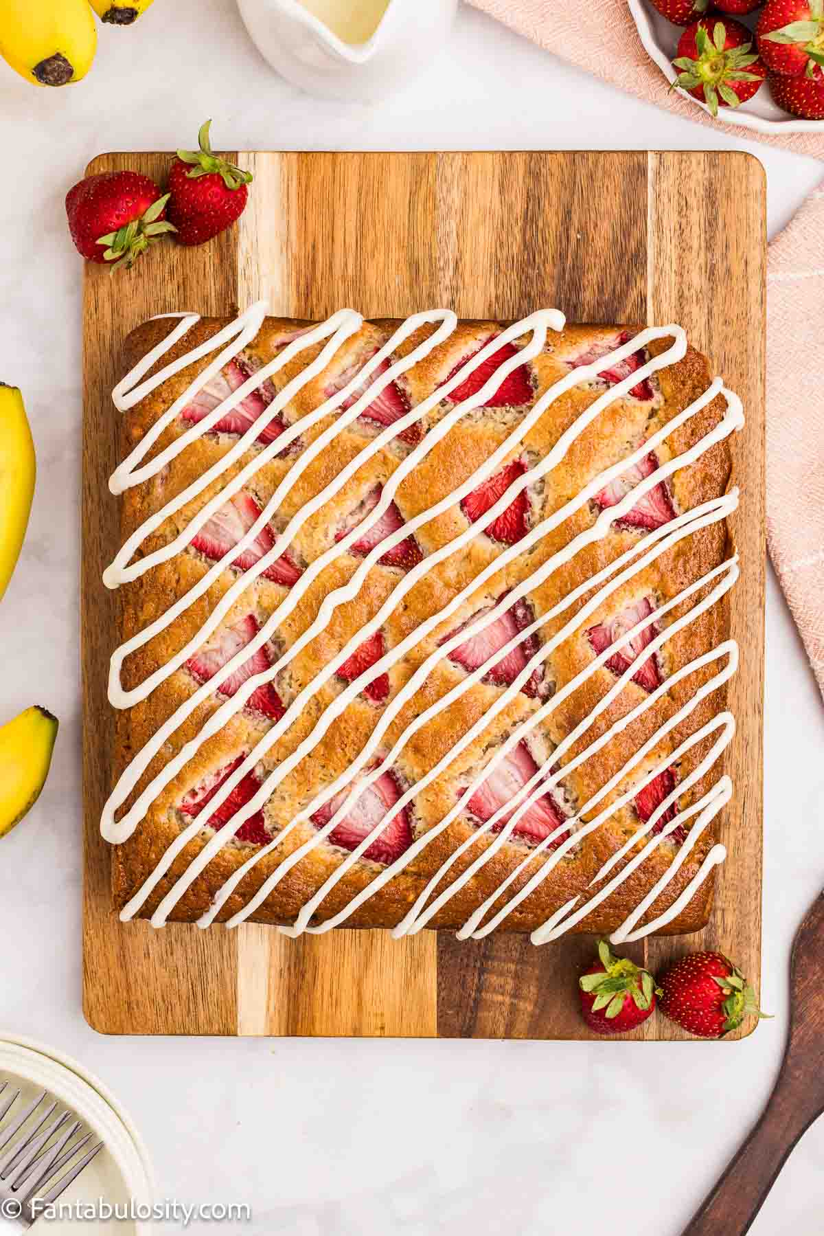 Strawberry banana cake topped with drizzle of cream cheese frosting, sitting on wooden cutting board.