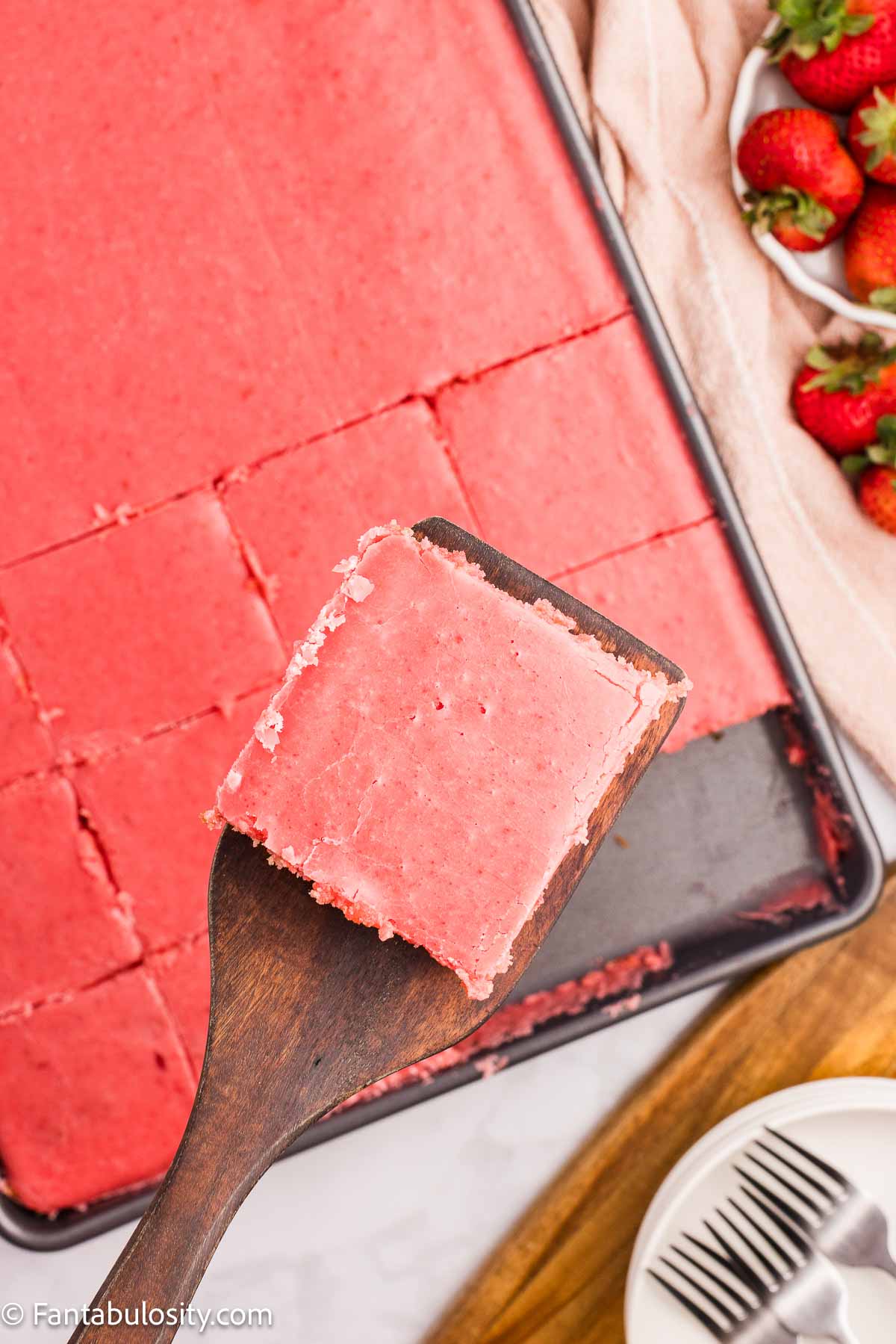 A slice of strawberry Texas sheet cake on a wooden spatula, above the rest of the cake.