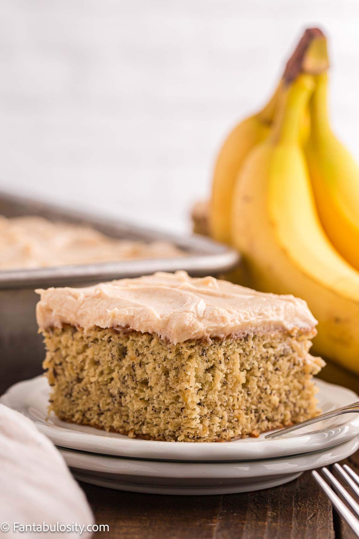 Slice of banana cake with brown butter frosting, sitting on round plate, next to bananas.