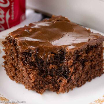 A slice of Coca-Cola cake on a vintage plate, sitting next to a Coca-Cola can.