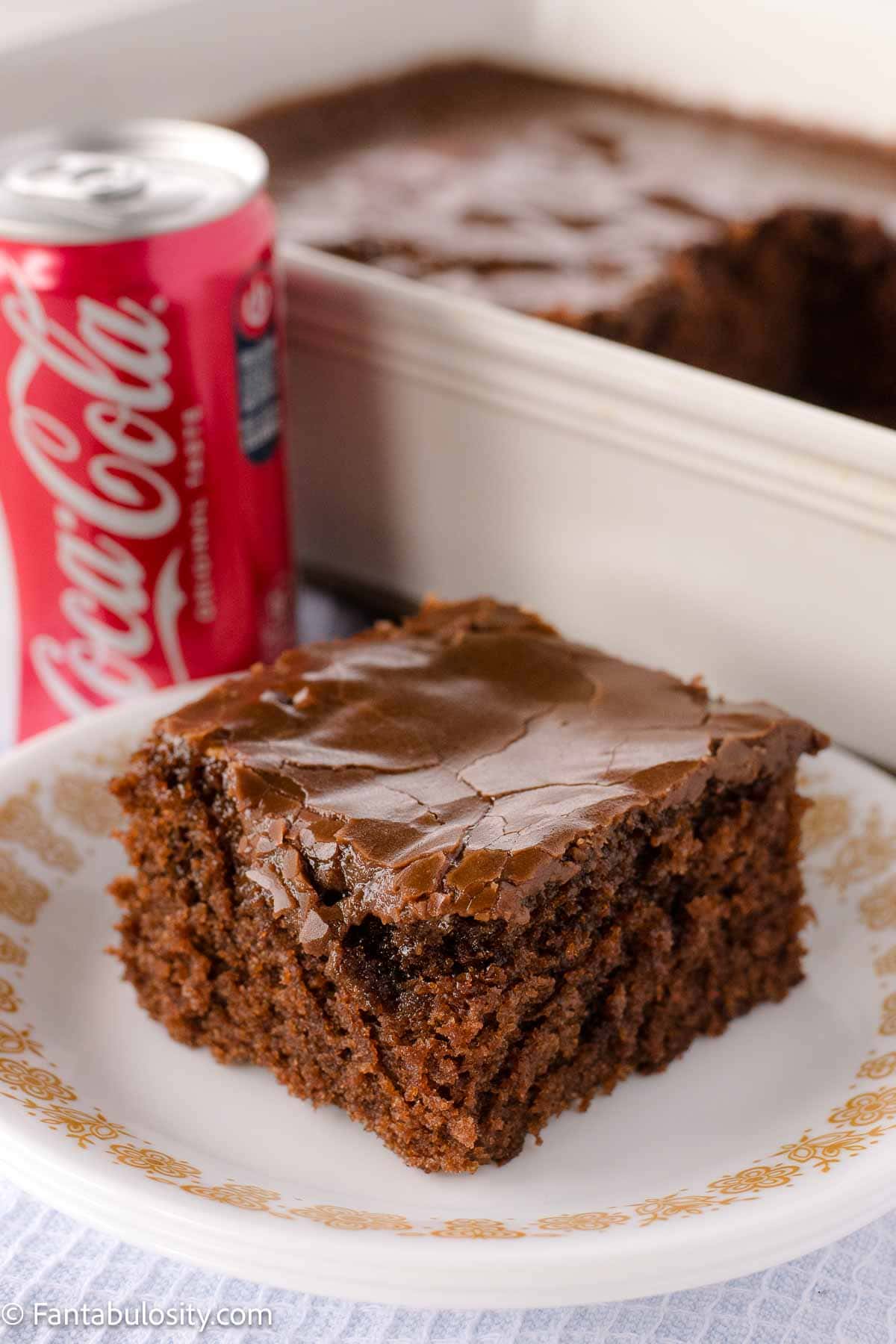 A slice of Coca-Cola cake on a vintage plate, sitting next to a can of Coca-Cola.
