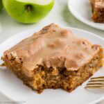 A square slice of apple dapple cake topped with caramel glaze on a white plate next to a green apple.