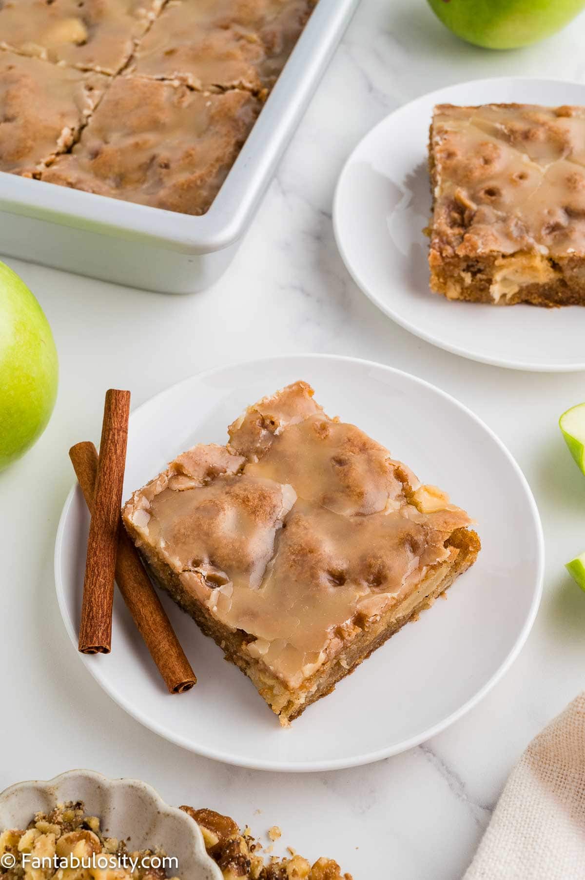 A slice of apple dapple cake on a white plate with cinnamon sticks, green apples, and the pan of cake behind it.