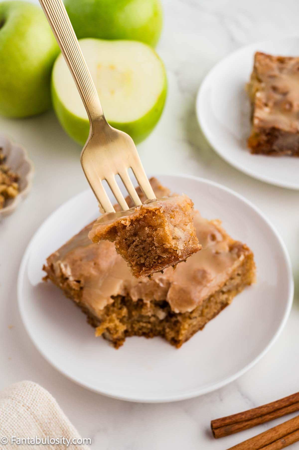 A gold fork lifting a bite from a slice of apple dapple cake on a white plate.