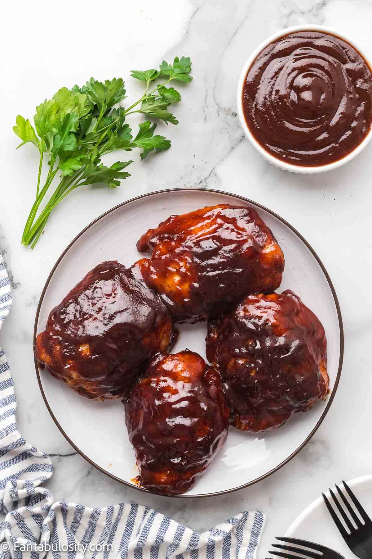 Baked BBQ chicken thighs on a white plate with a bowl of BBQ sauce and fresh parsley.