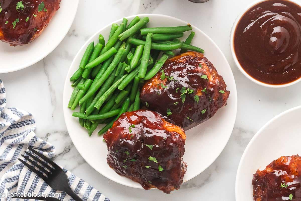 Baked BBQ chicken thighs served on a white plate with green beans and a bowl of BBQ sauce.
