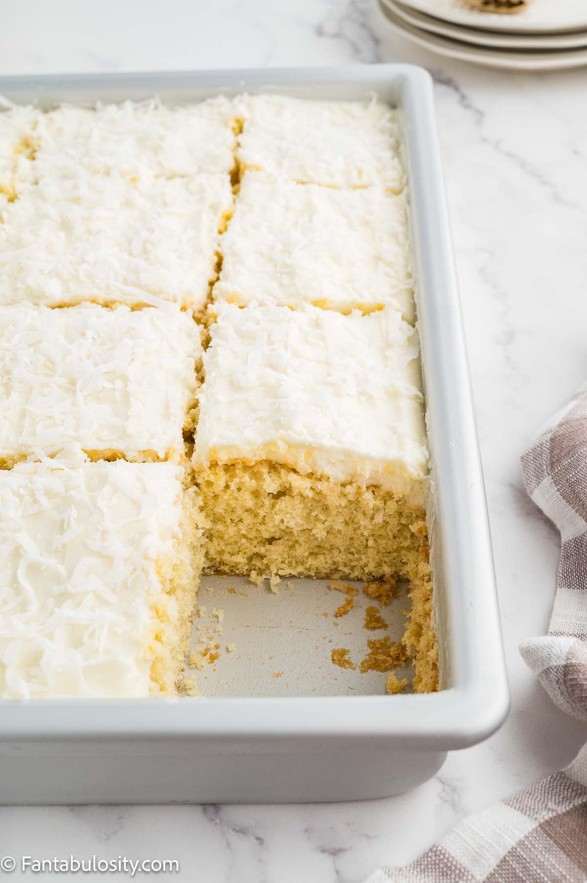 Coconut sheet cake with coconut frosting, sliced in the baking pan with one piece lifted out.