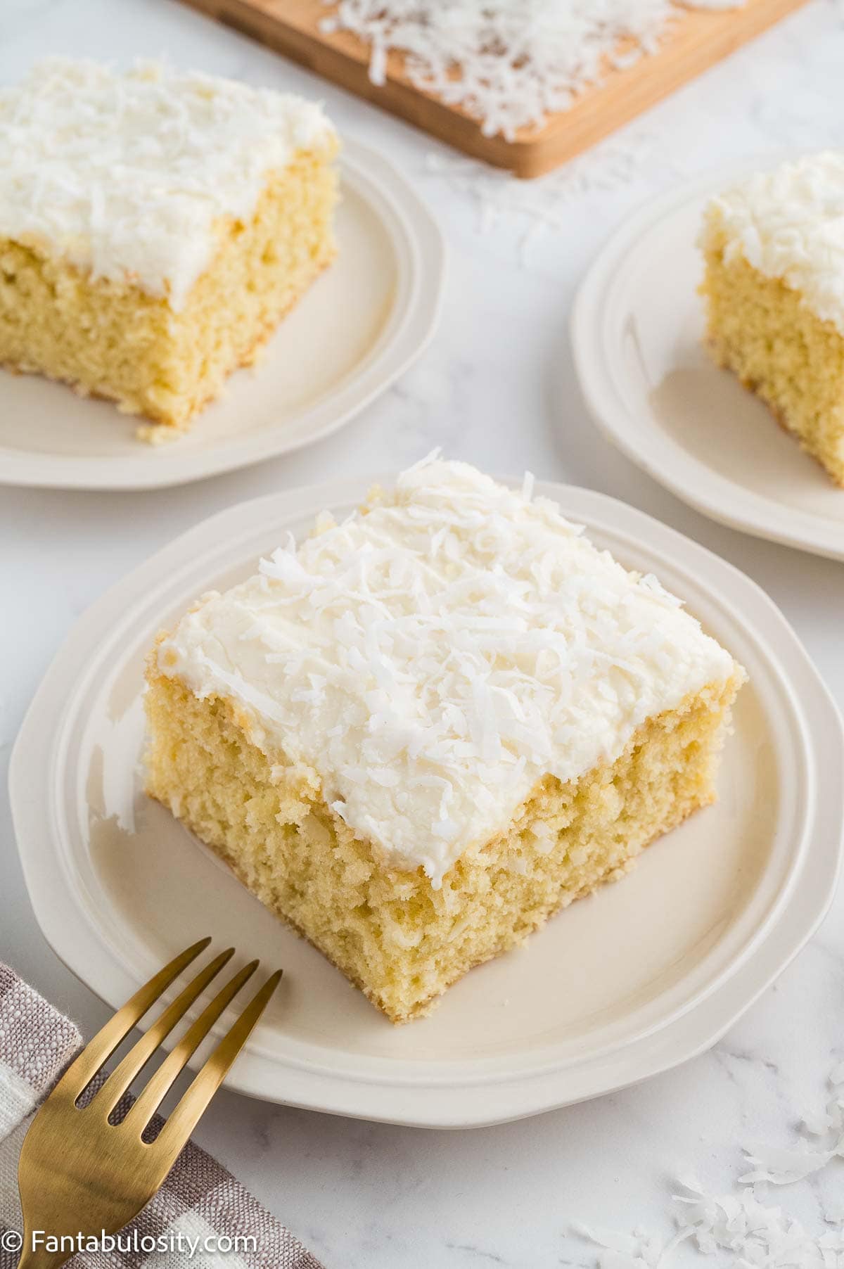 Two slices of coconut sheet cake on white plates with a gold fork.