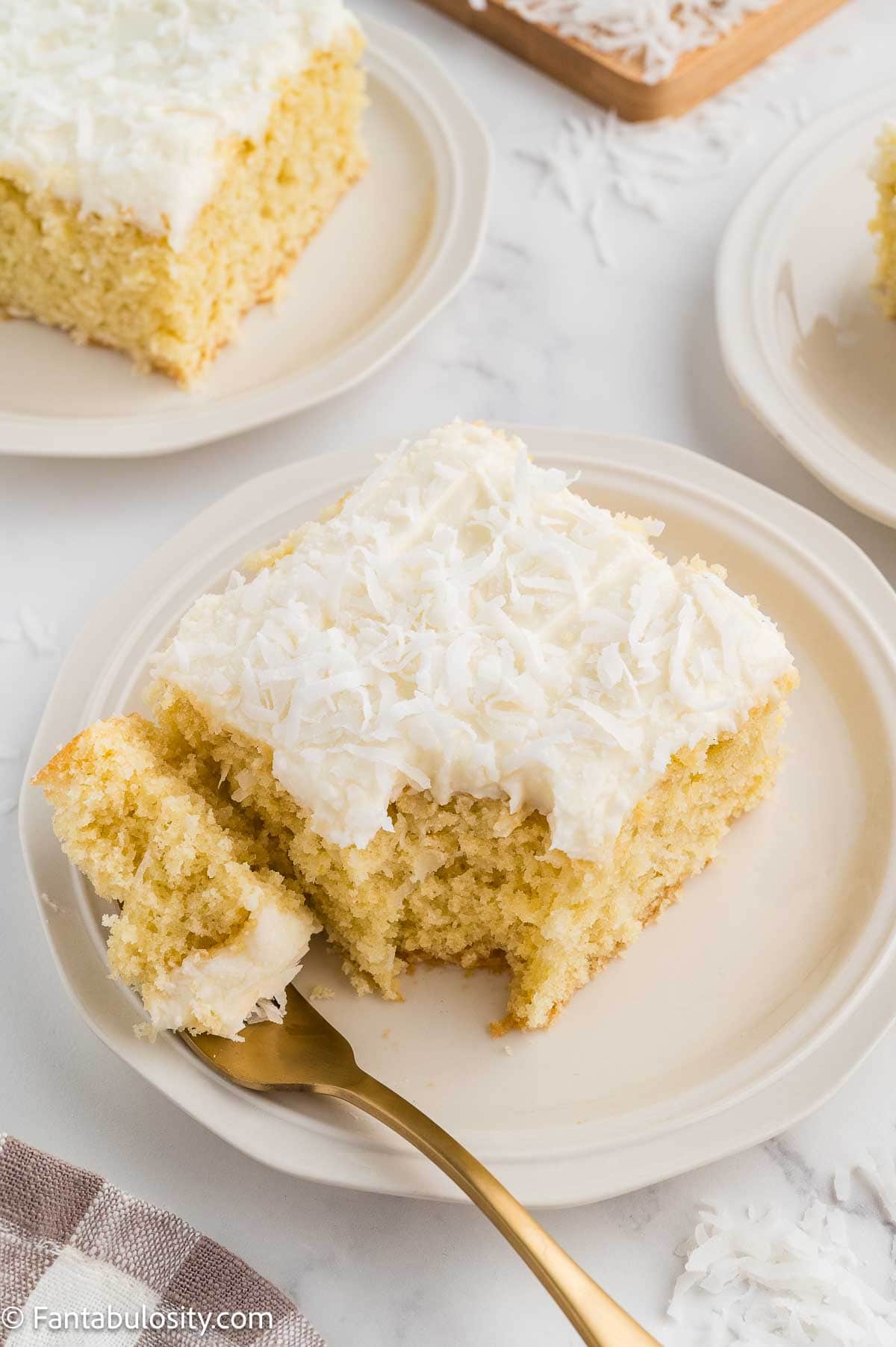 Slice of coconut sheet cake on a white plate with a bite taken out.