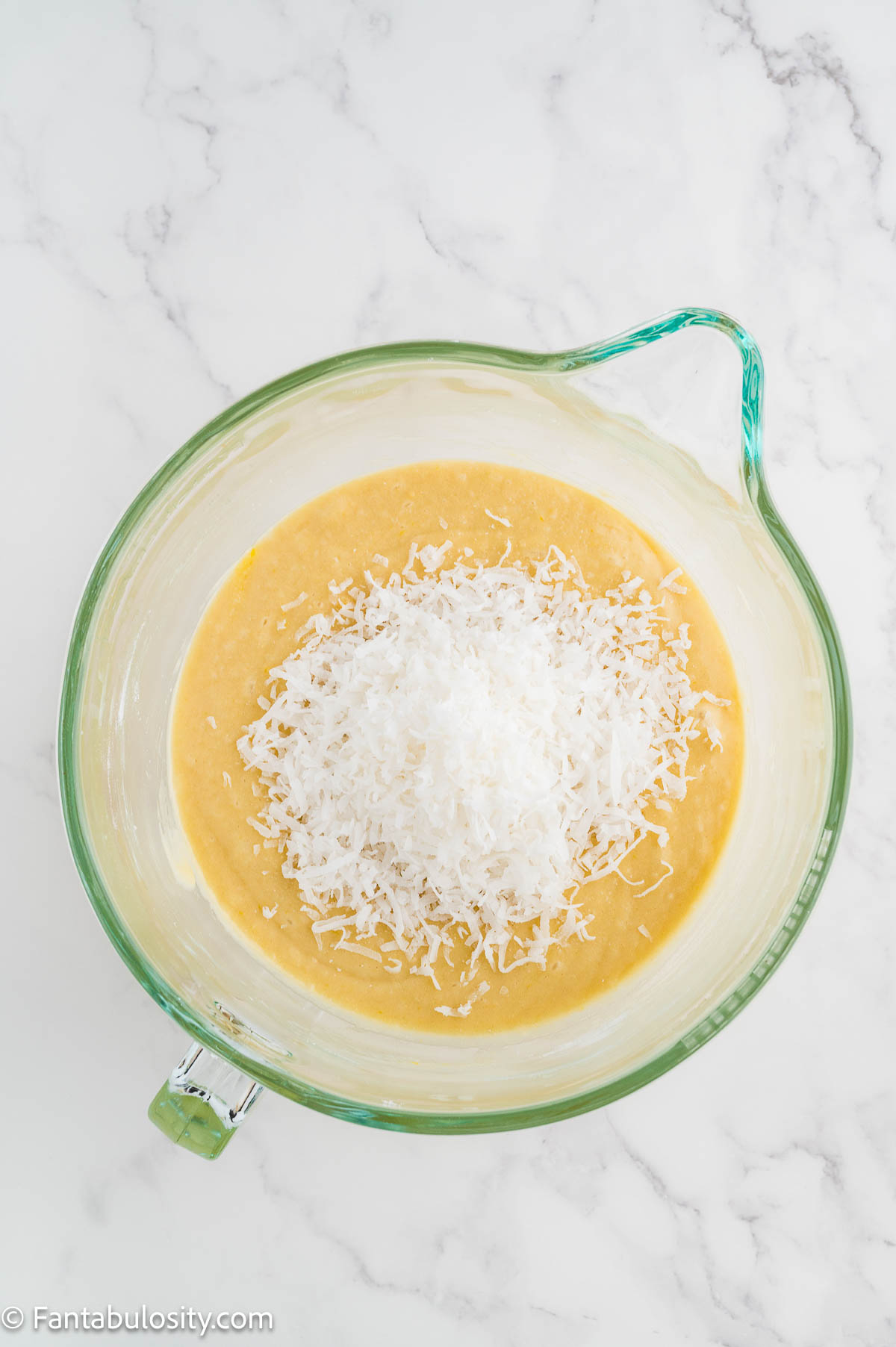 Shredded coconut and dry ingredients added to the cake batter in a mixing bowl.