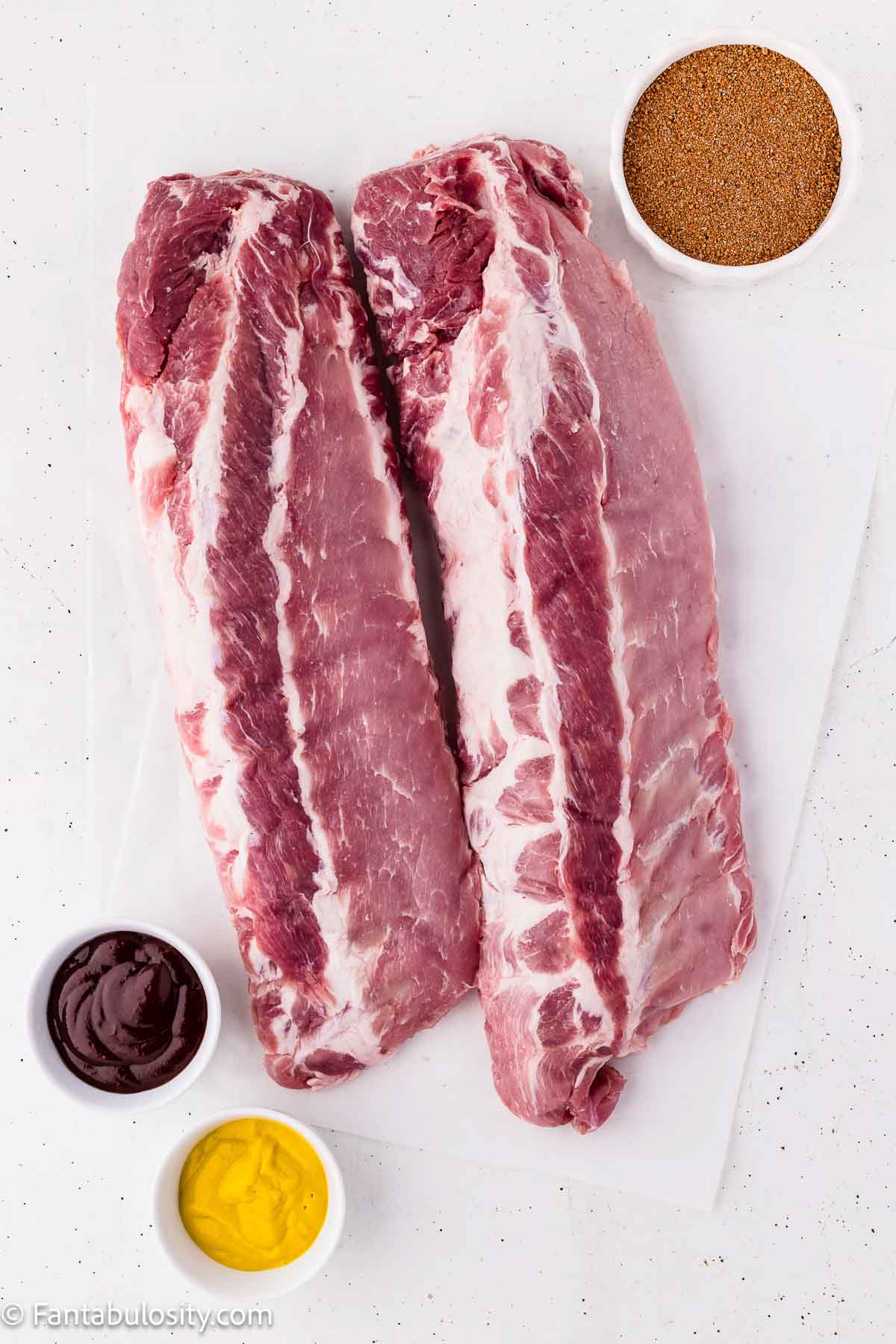 Two racks of raw baby back ribs with bowls of dry rub, yellow mustard, and BBQ sauce