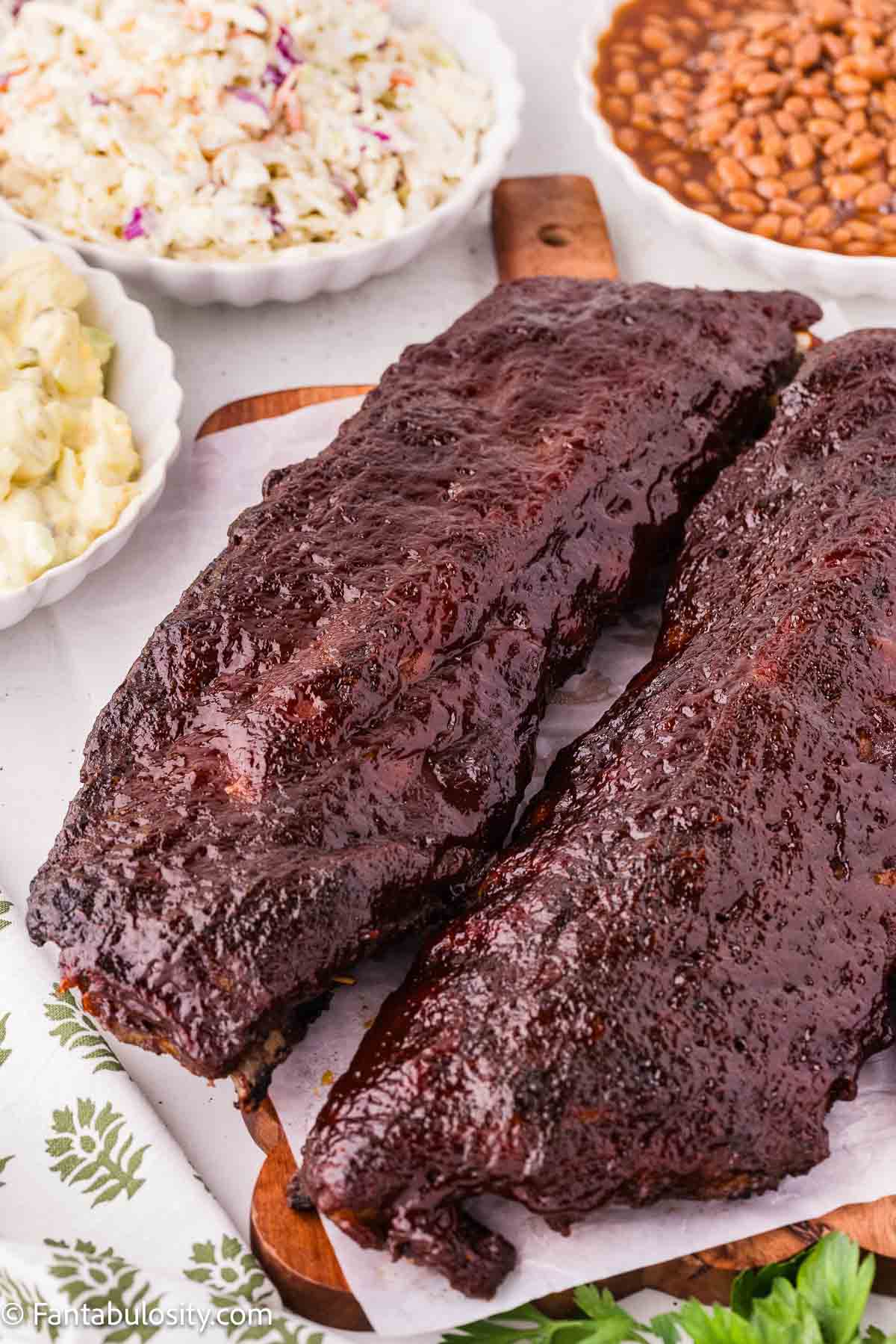 A rack of oven baked ribs on a wooden board with coleslaw, baked beans, and potato salad