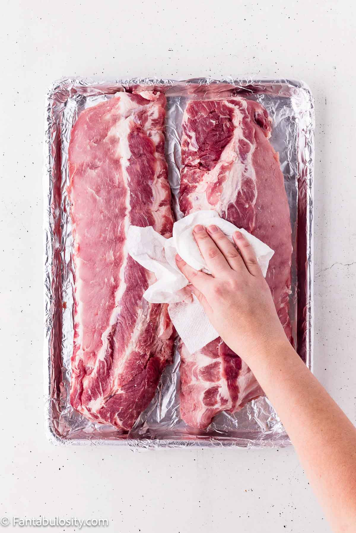 Blotting raw baby back ribs dry with a paper towel on a foil-lined sheet pan