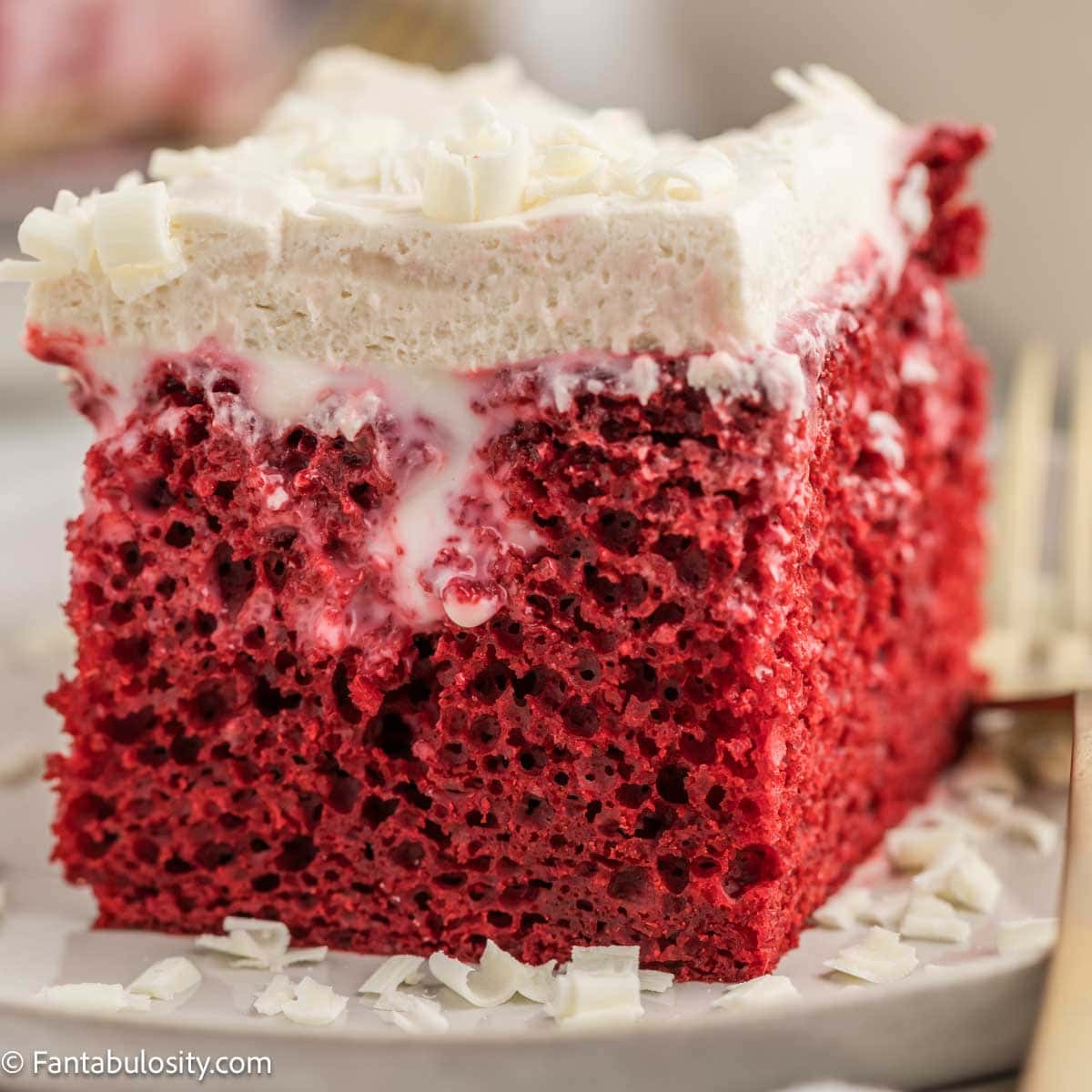 Red velvet poke cake slice with cream cheese frosting and white chocolate curls on a plate.
