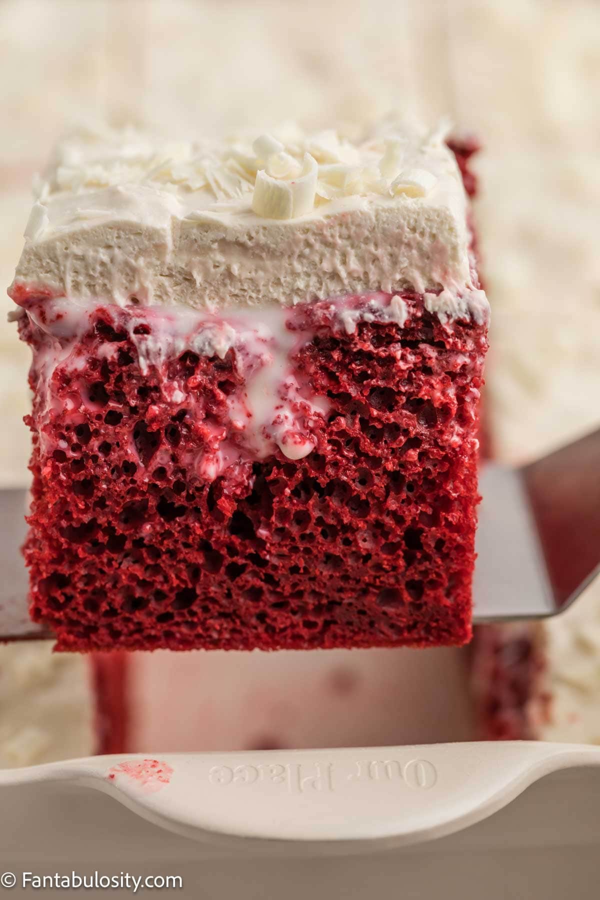 A slice of red velvet poke cake lifted on a cake server showing the filling soaked into the red cake.