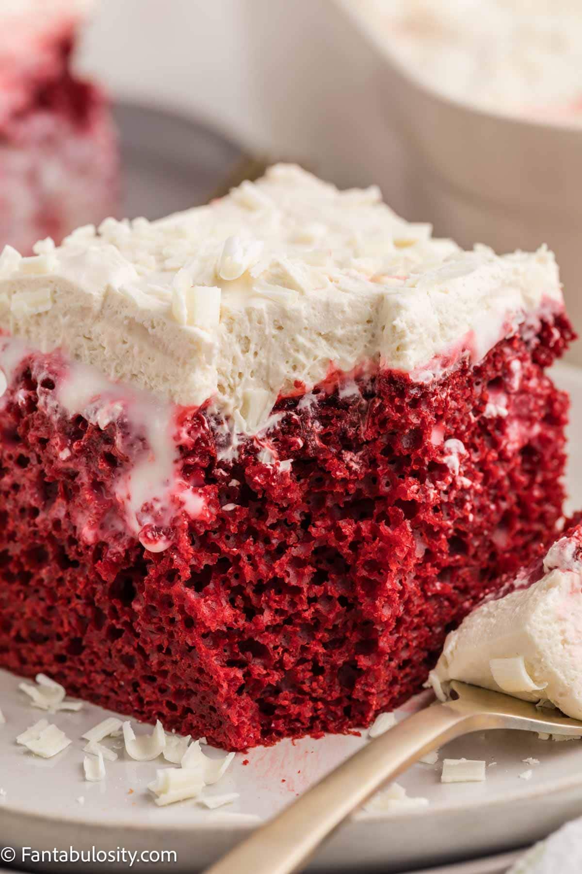 A slice of red velvet poke cake on a plate with white chocolate shavings and filling soaked through the cake.