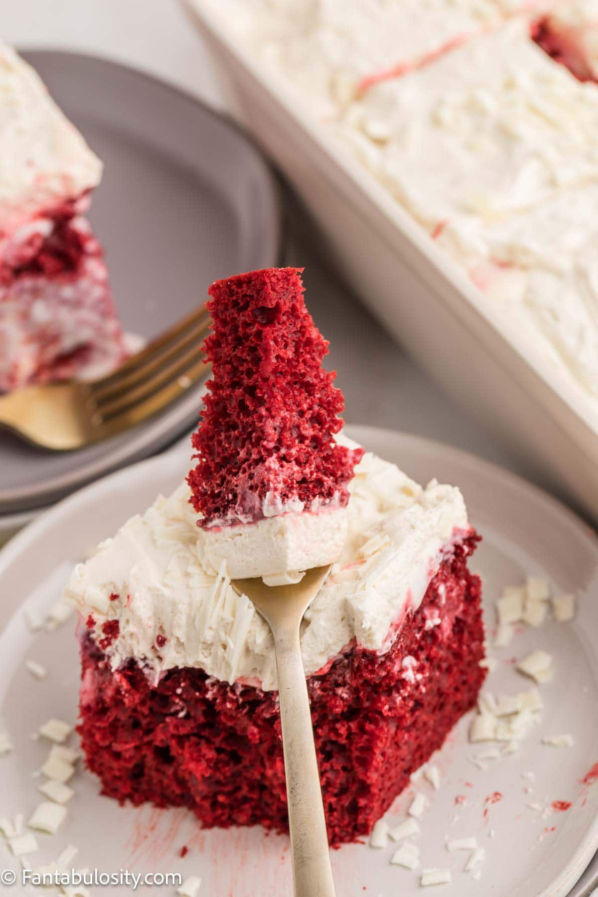 A fork taking a bite from a slice of red velvet poke cake on a plate.