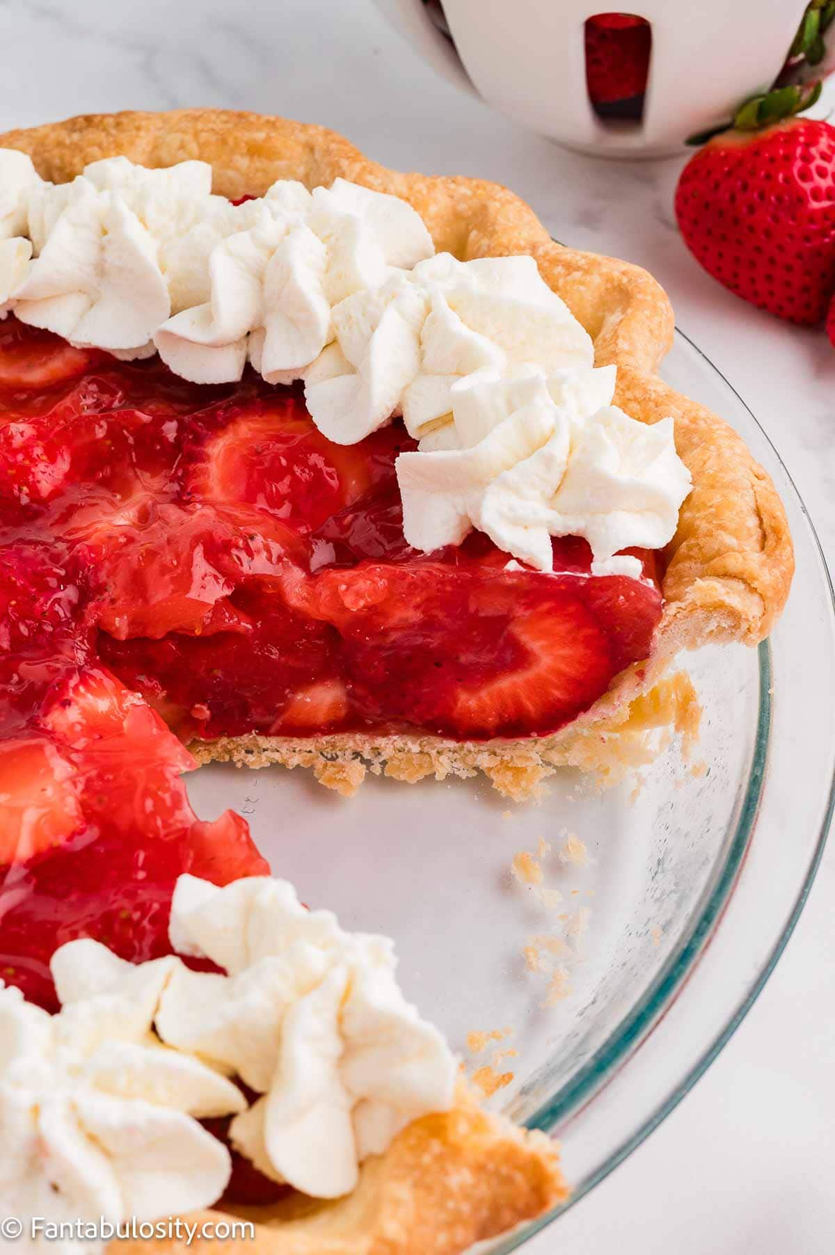 Close-up of fresh strawberry pie with a slice cut out, showing whole strawberries in a glossy red glaze under piped whipped cream.