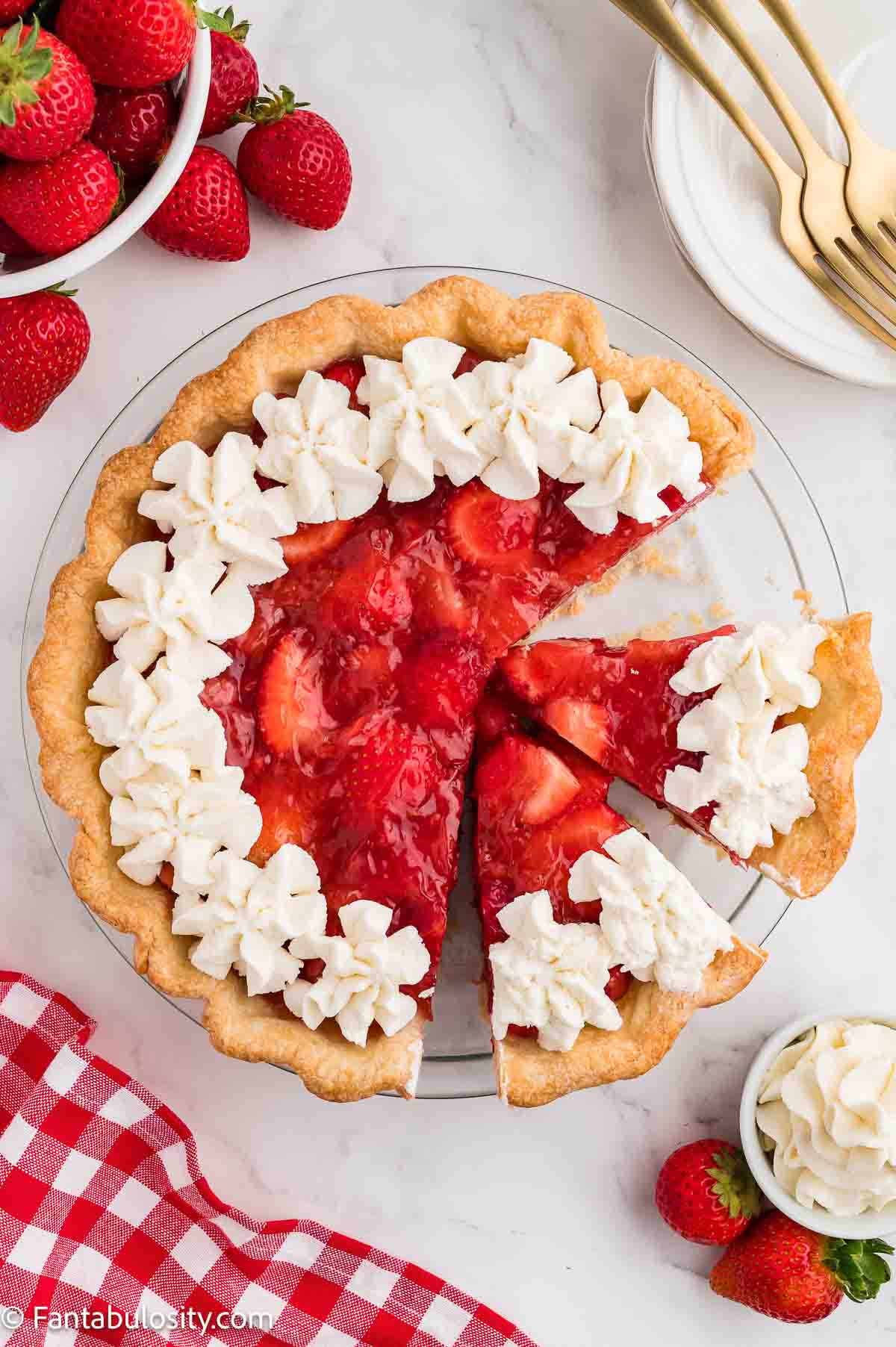 Overhead view of a fresh strawberry pie with whipped cream around the edge and one slice pulled away.