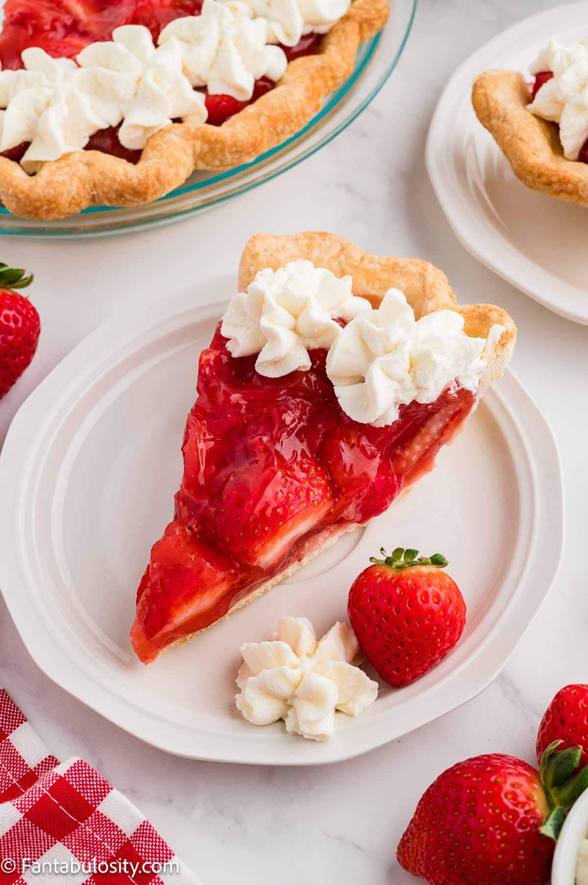 A slice of fresh strawberry pie on a white plate with whipped cream and the rest of the pie behind it.