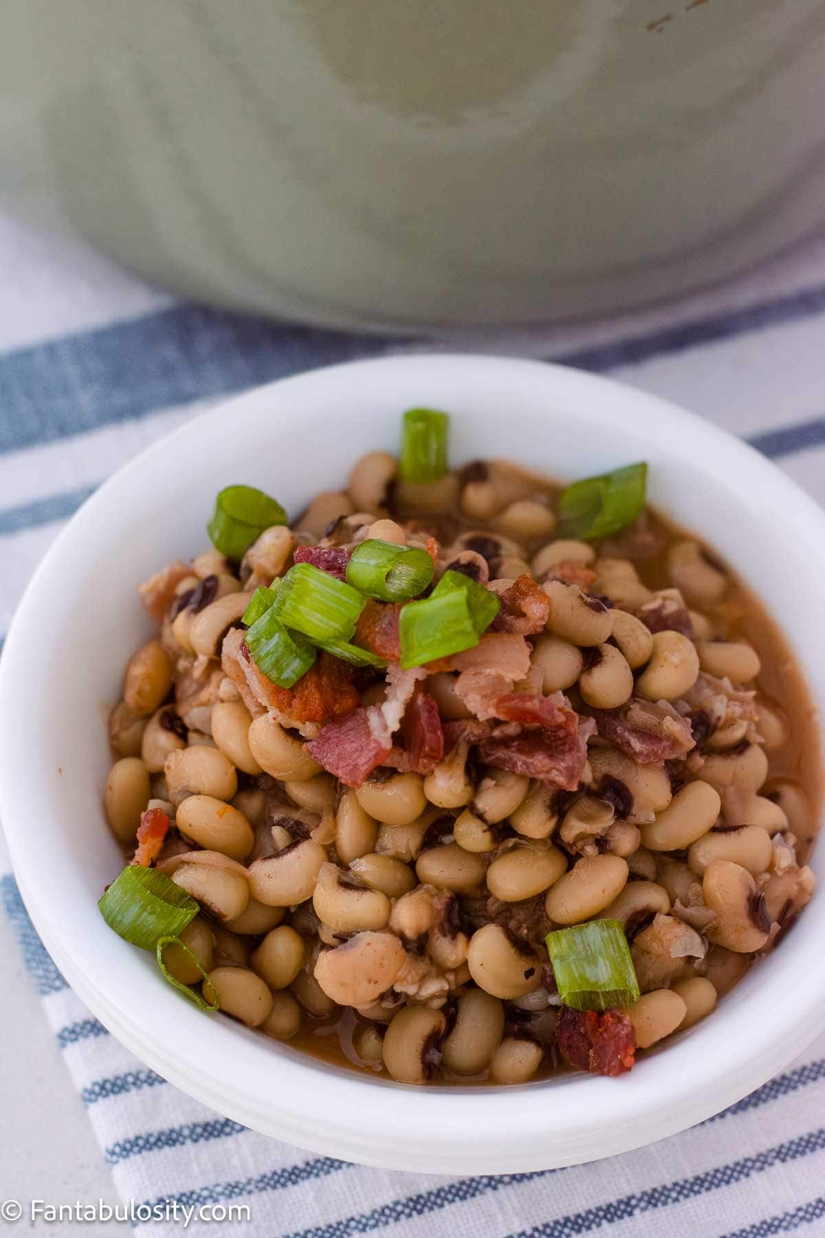 A bowl of black eyed peas topped with bacon and green onions on a striped towel.