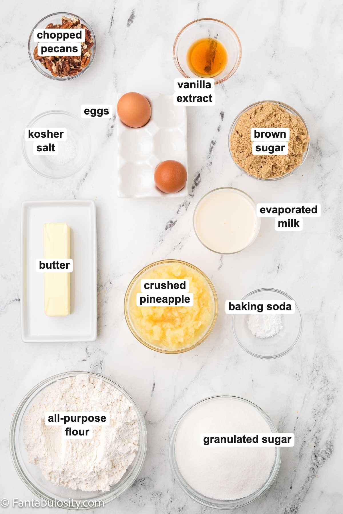 Labeled ingredients for granny cake on a marble counter: chopped pecans, vanilla extract, kosher salt, eggs, brown sugar, evaporated milk, butter, crushed pineapple, baking soda, all-purpose flour, and granulated sugar.