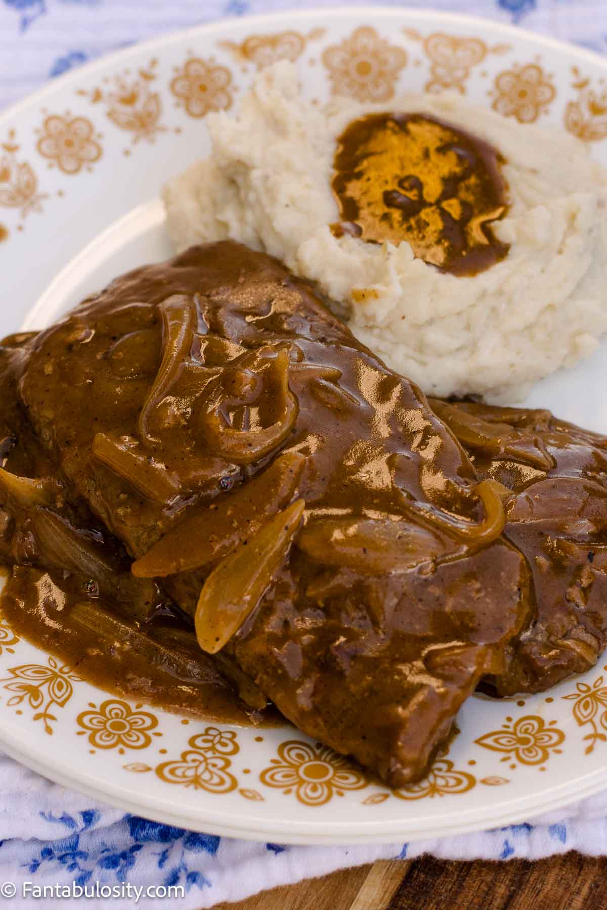 Cube steak covered in brown onion gravy with mashed potatoes on a floral plate.