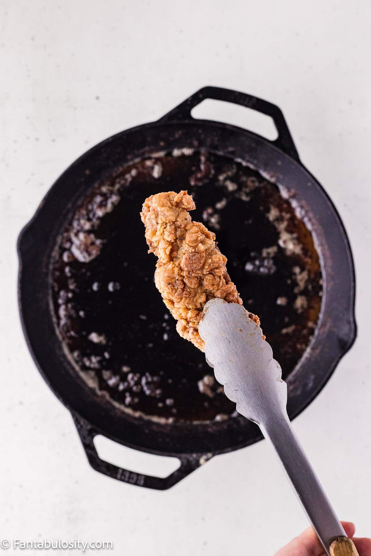 A golden steak finger lifted from hot oil in a cast-iron skillet.