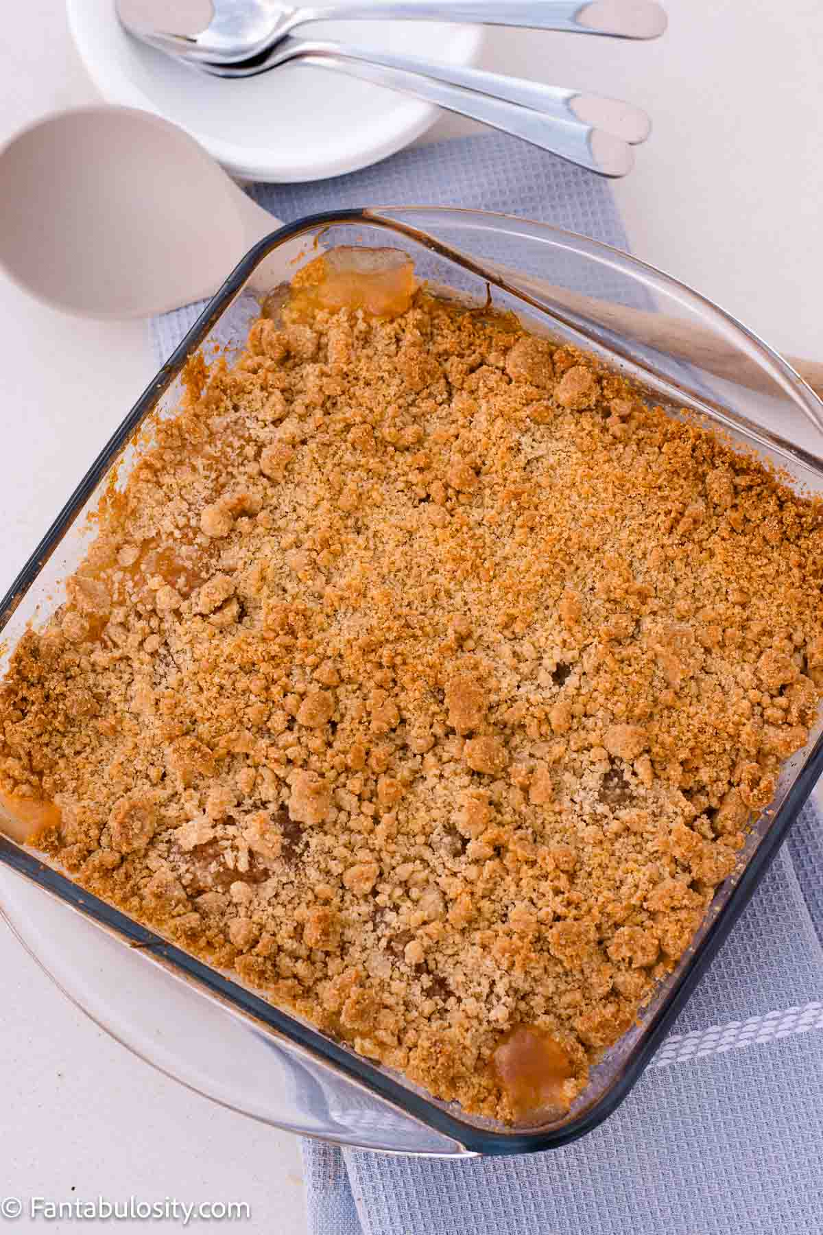 Golden baked apple crumble in a square glass dish with apple filling visible around the edges.