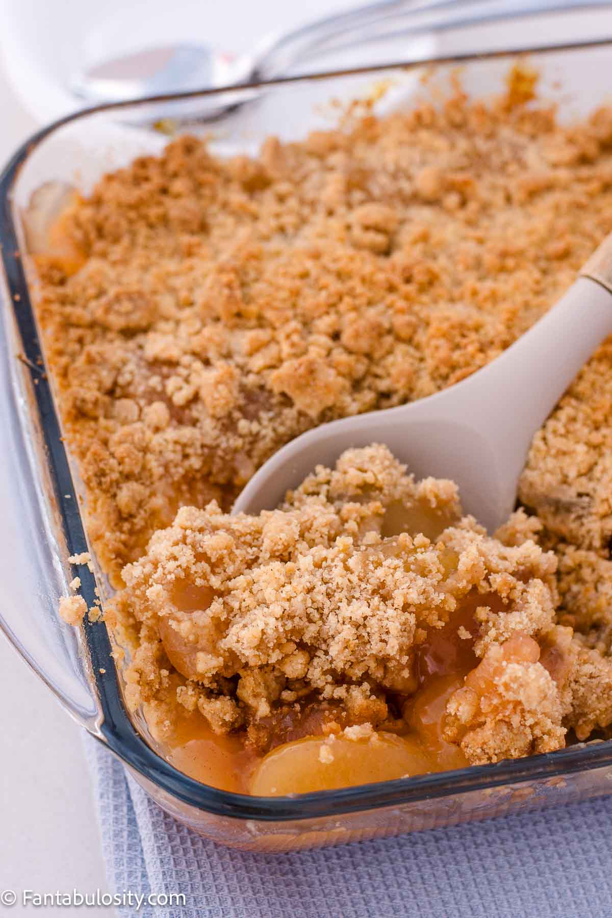 A spoon lifting apple filling and golden crumble topping from a glass baking dish.