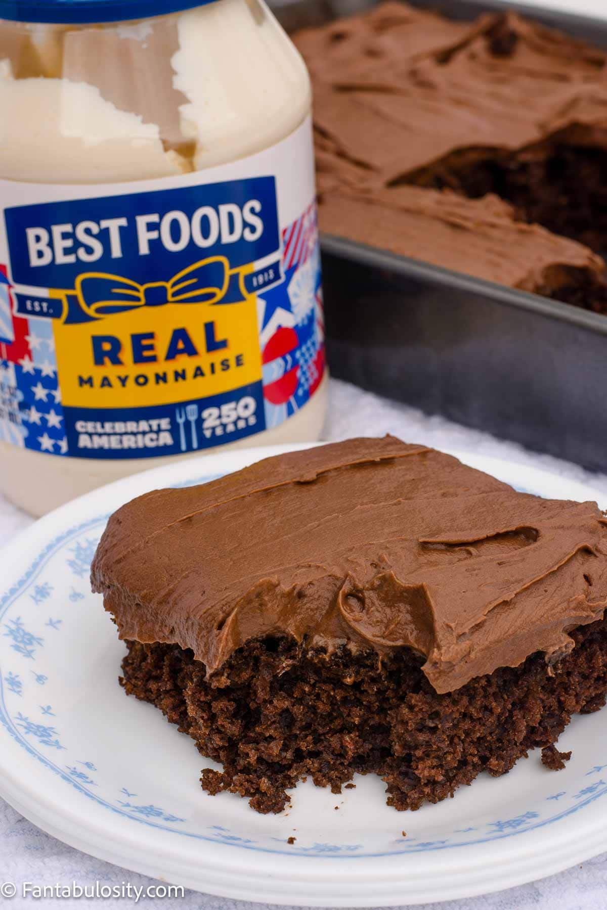 Slice of chocolate mayonnaise cake on a plate with a jar of mayonnaise and the pan of cake in the background.
