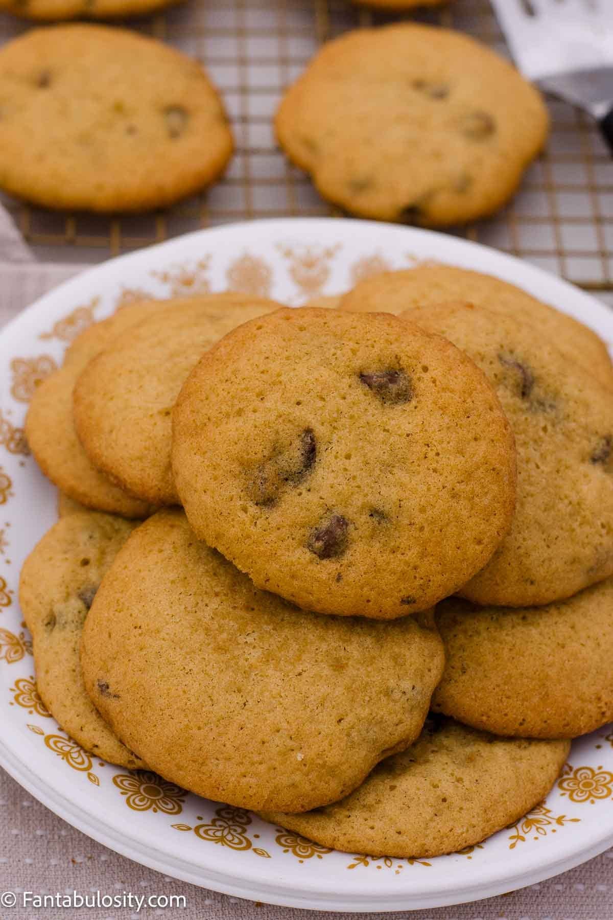 A plate of golden chocolate chip cookies with more cookies cooling on a wire rack behind it.