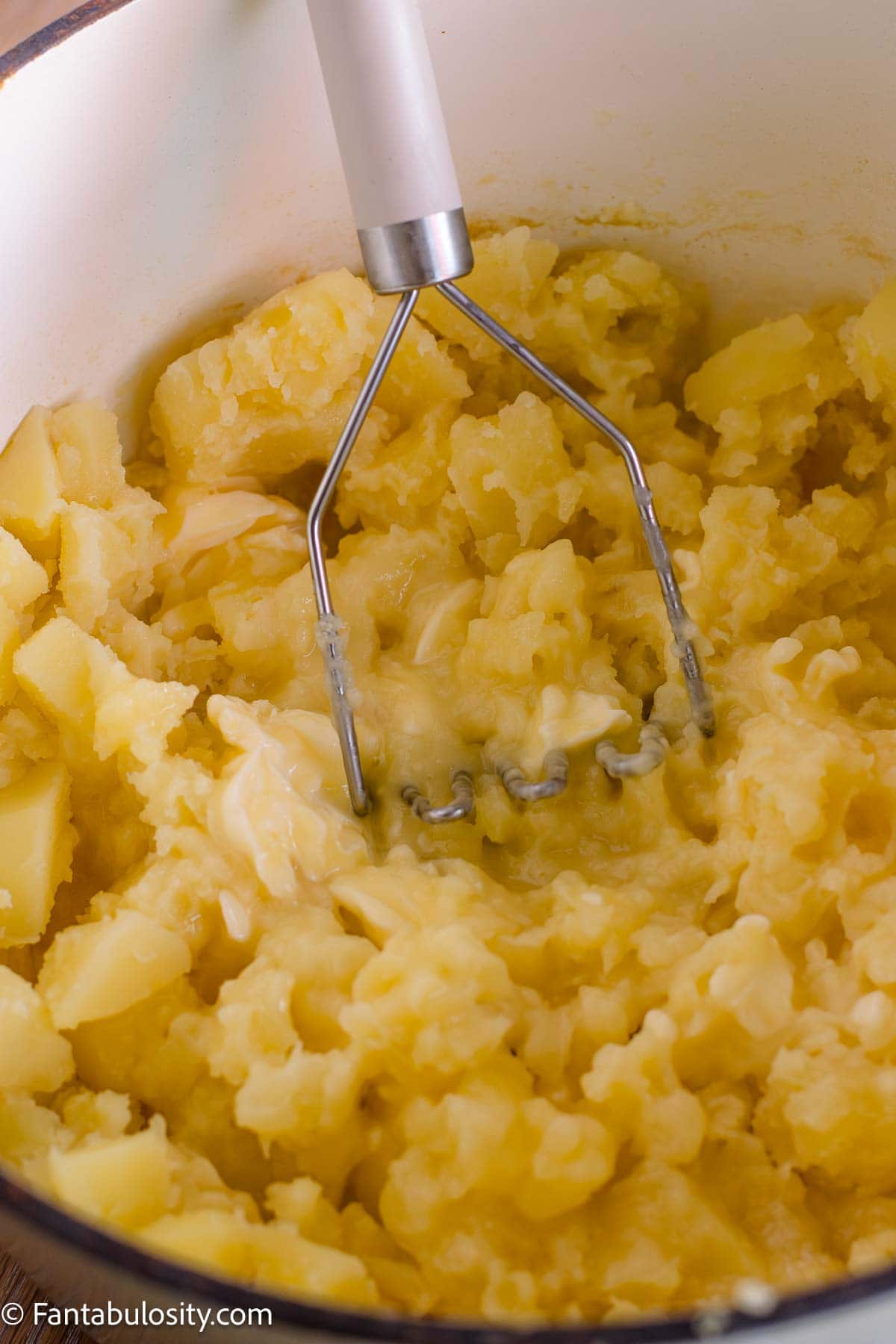 A potato masher working butter into hot cooked potatoes.